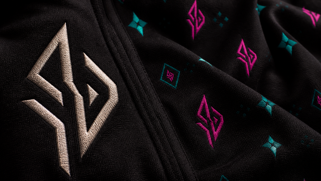 Gesticktes SUÐR-Emblem auf schwarzem Stoff, daneben dasselbe Zeichen als Monogramm-Muster in Pink und Türkis
