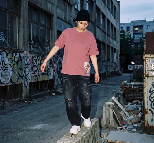 SUÐR REBELLION – Oversized Tee in Altrosa mit kleinem Frontprint und SUÐR-Logo am Ärmel, fotografiert in einer Graffiti-Gasse
